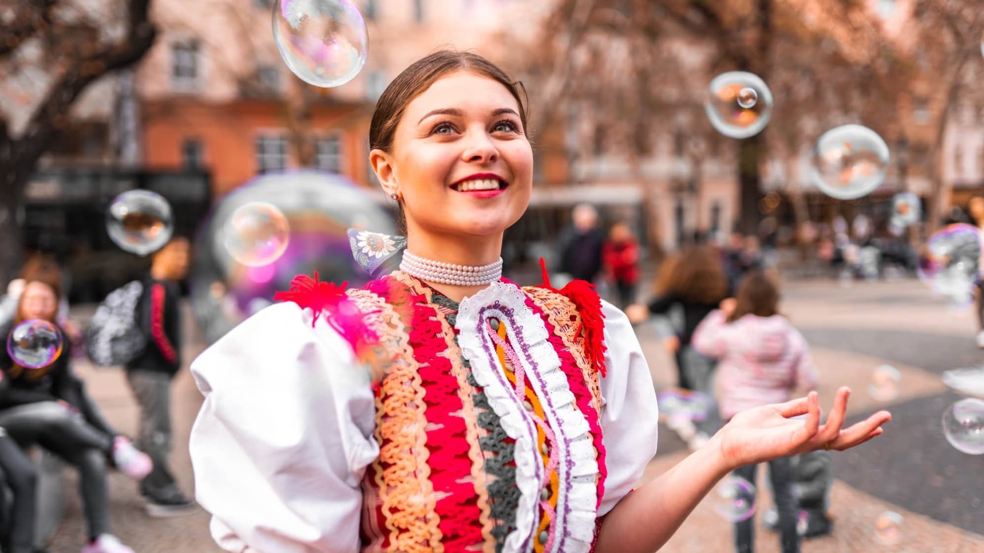 A dancer in an embroidered folk costume laughing among soap bubbles on a city square