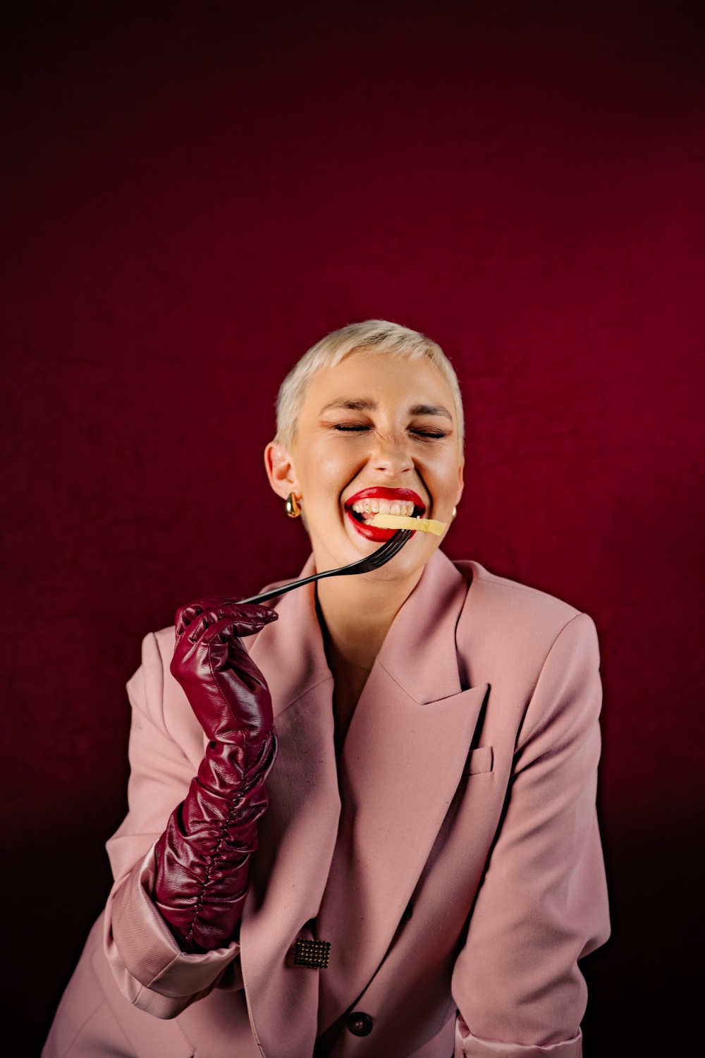Sofia Vrabčeková in a pink blazer and burgundy leather glove, eating off a fork against deep red