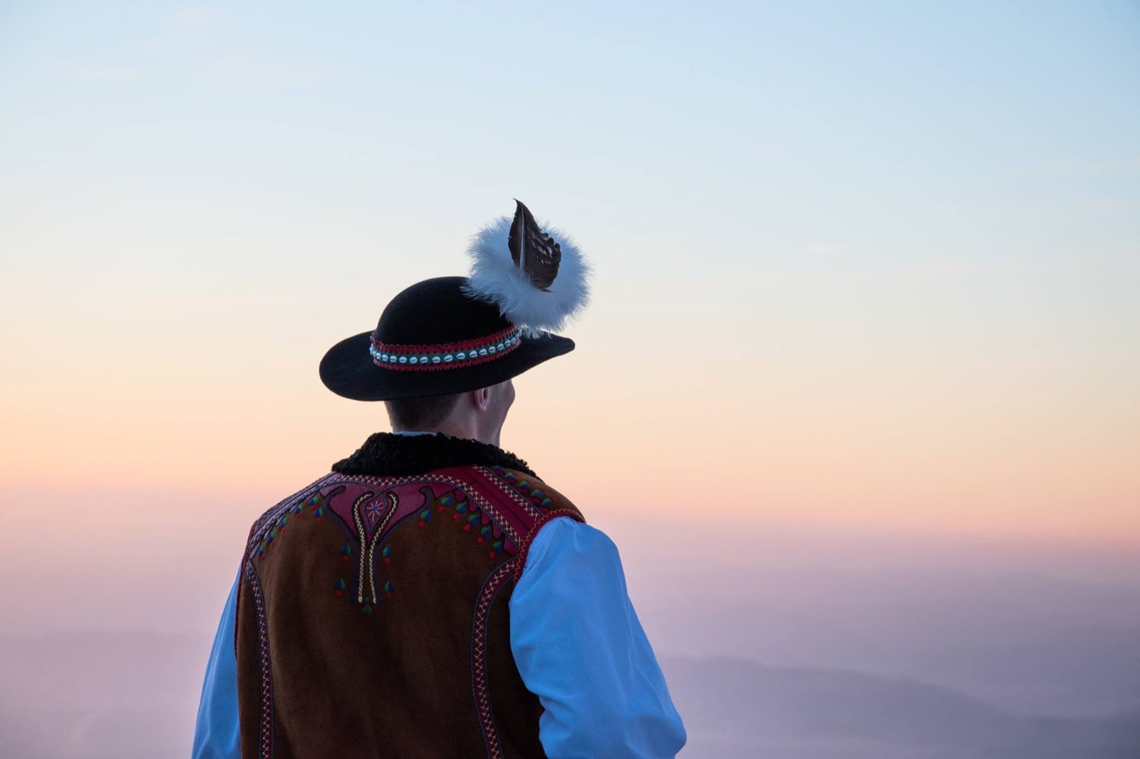 A dancer in an embroidered vest and feathered hat seen from behind, looking out over a valley at sunrise